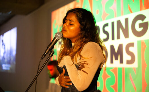 a brown woman with her eyes closed signing into a microphone in front of a projected graphic that reads Rehearsing Freedoms slightly obscured