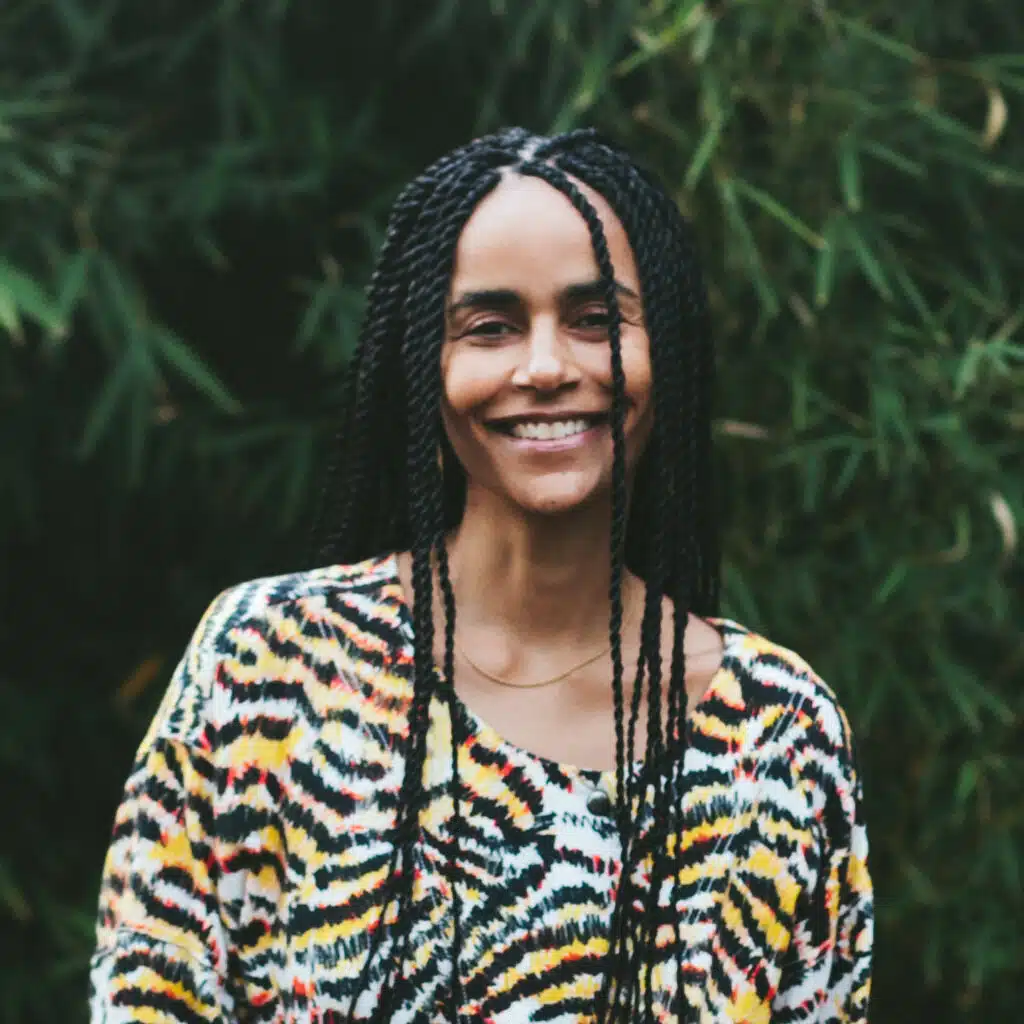 Nkem Ndefo, a Black woman with long black twists smiling at the camera weraing a mixed animal print shirt in front of greenery in the background