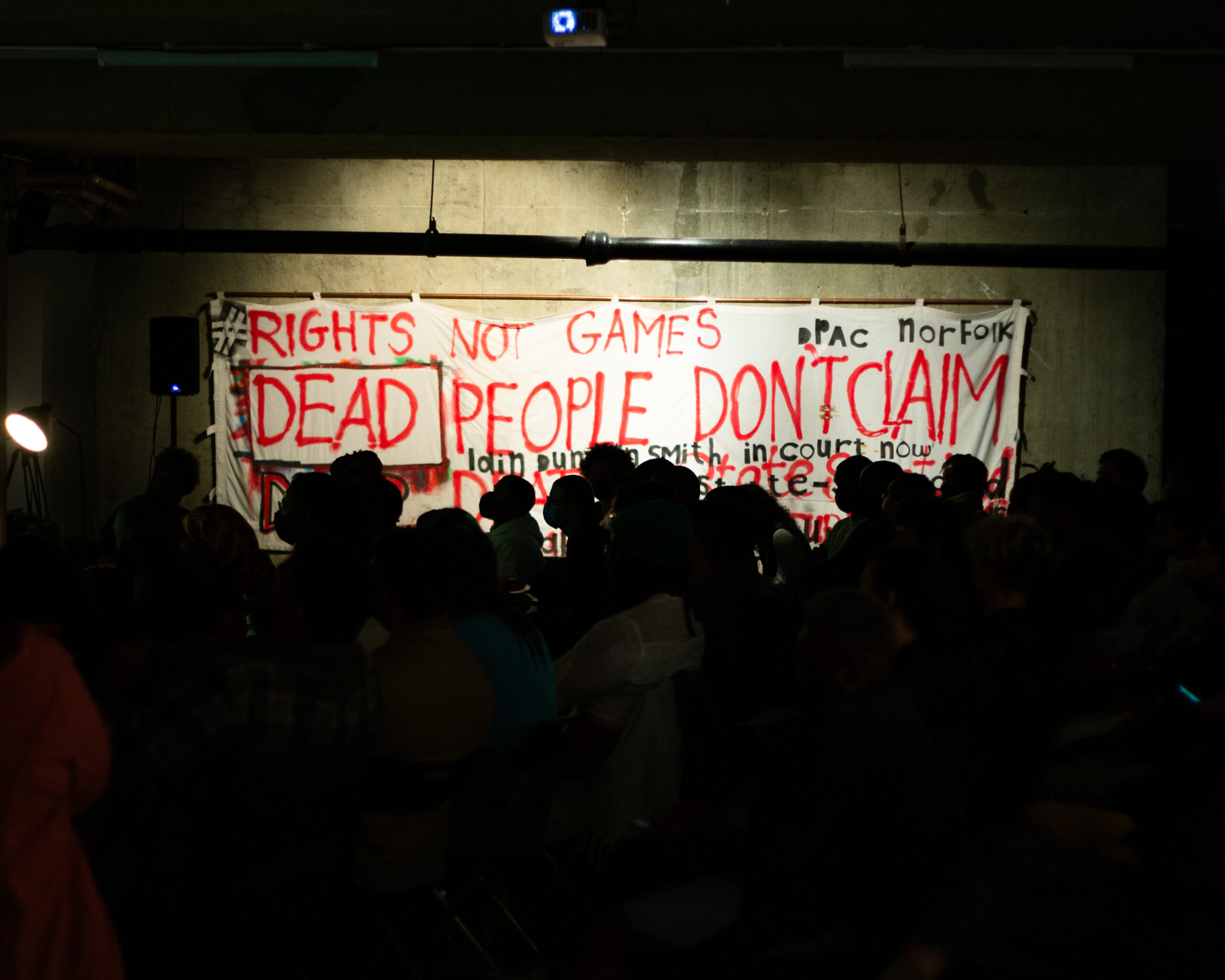 a crowd is in shadow in the foreground and a banner in the background reads 'dead people don't claim'