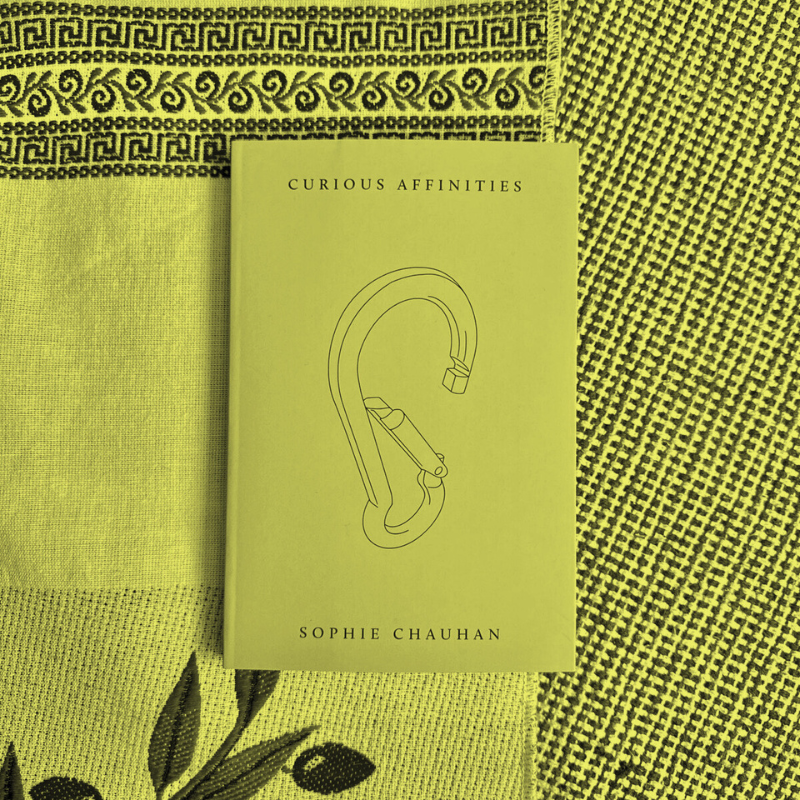 front cover of Sophie Chauhan's book Curious Affinities which has a mustard yellow cover and is sitting on top of a woven blanket. Image has yellow-green filter on it