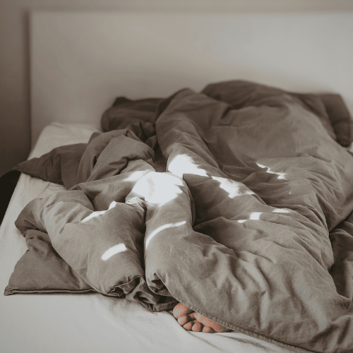 A sun streaked brown-duvet crumpled up on a white bed with two feet just peeking out at the bottom