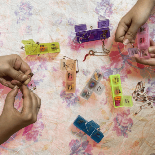 Multicoloured pill boxes and safety pins are spread over a flowery surface with hands holding and playing with the boxes and pins