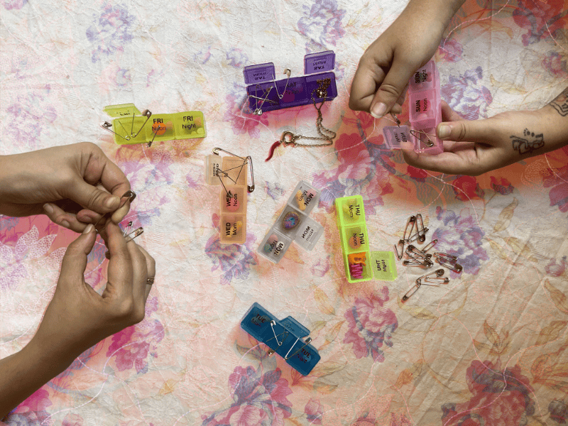 Multicoloured pill boxes and safety pins are spread over a flowery surface with hands holding and playing with the boxes and pins