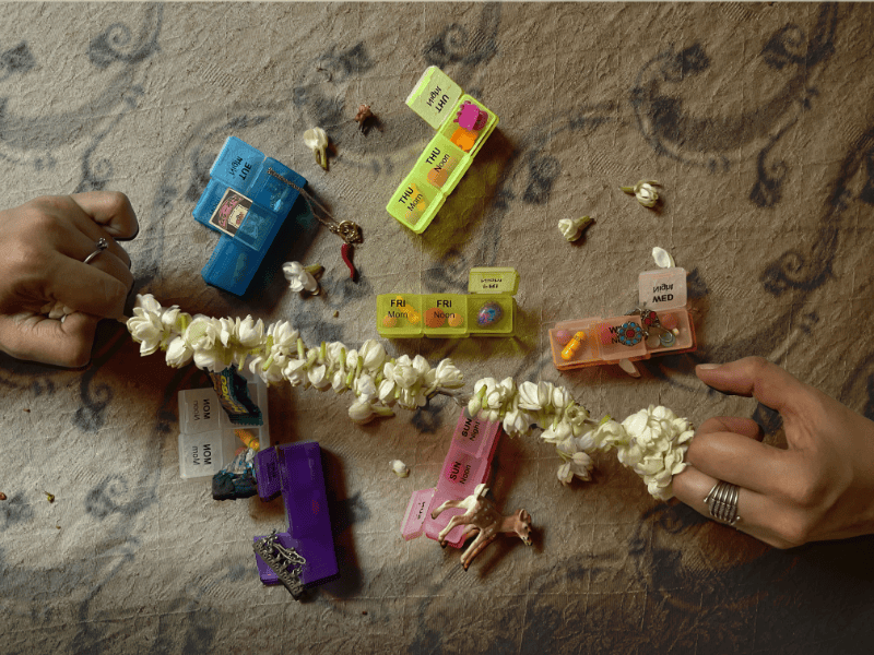 Colourful pill boxes are spread out on a grey decorated surface with a string of white flowers being held across the top by two hands grasping each end in their fingers