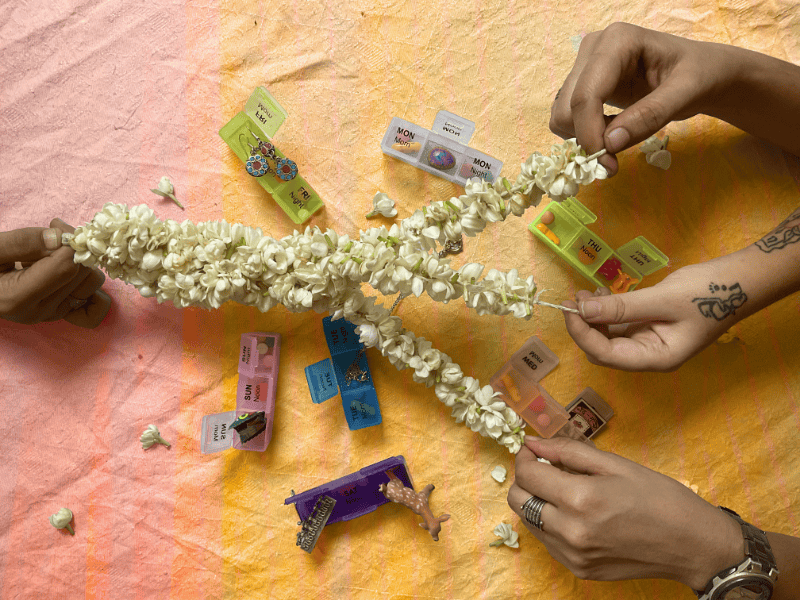 Colourful pill boxes are spread out on an orange and pink surface with multiple strings of white flowers being held across the top by various hands grasping each end in their fingers