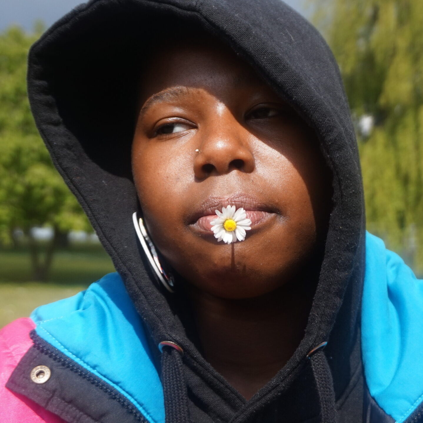 Aiyana - a Black young person - wears a black hoodie, a bright pink and blue jacket, and large Africa shaped earrings. They are outside surrounded by out-of-focus trees. They have a daisy flower in their mouth and look past the camera into the distance.